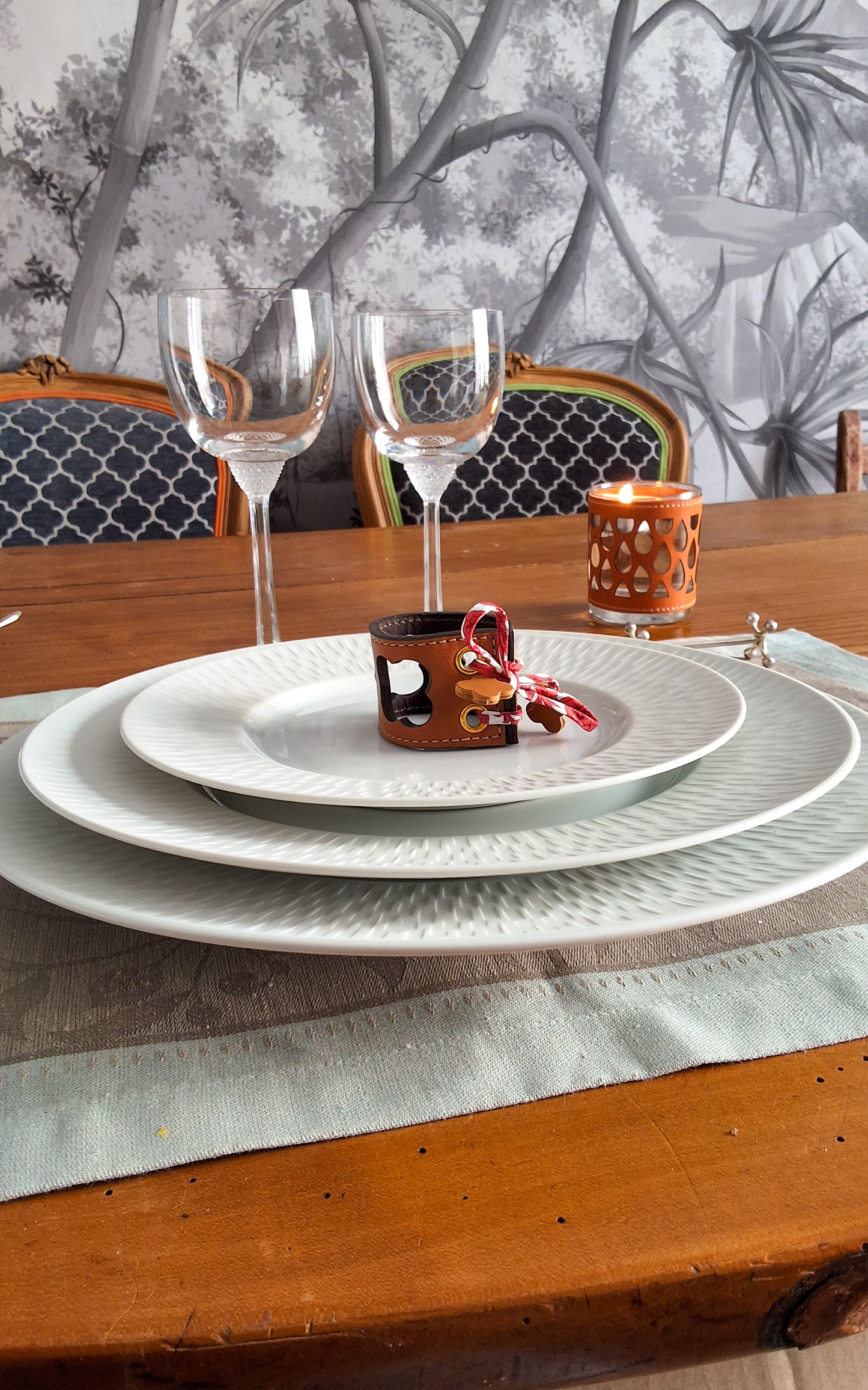 Table élégante dressee avec 3 assietes les unes sur les autres deux verresa pied et le rond de serviette en cuir trefle au centre de la derniere assiette