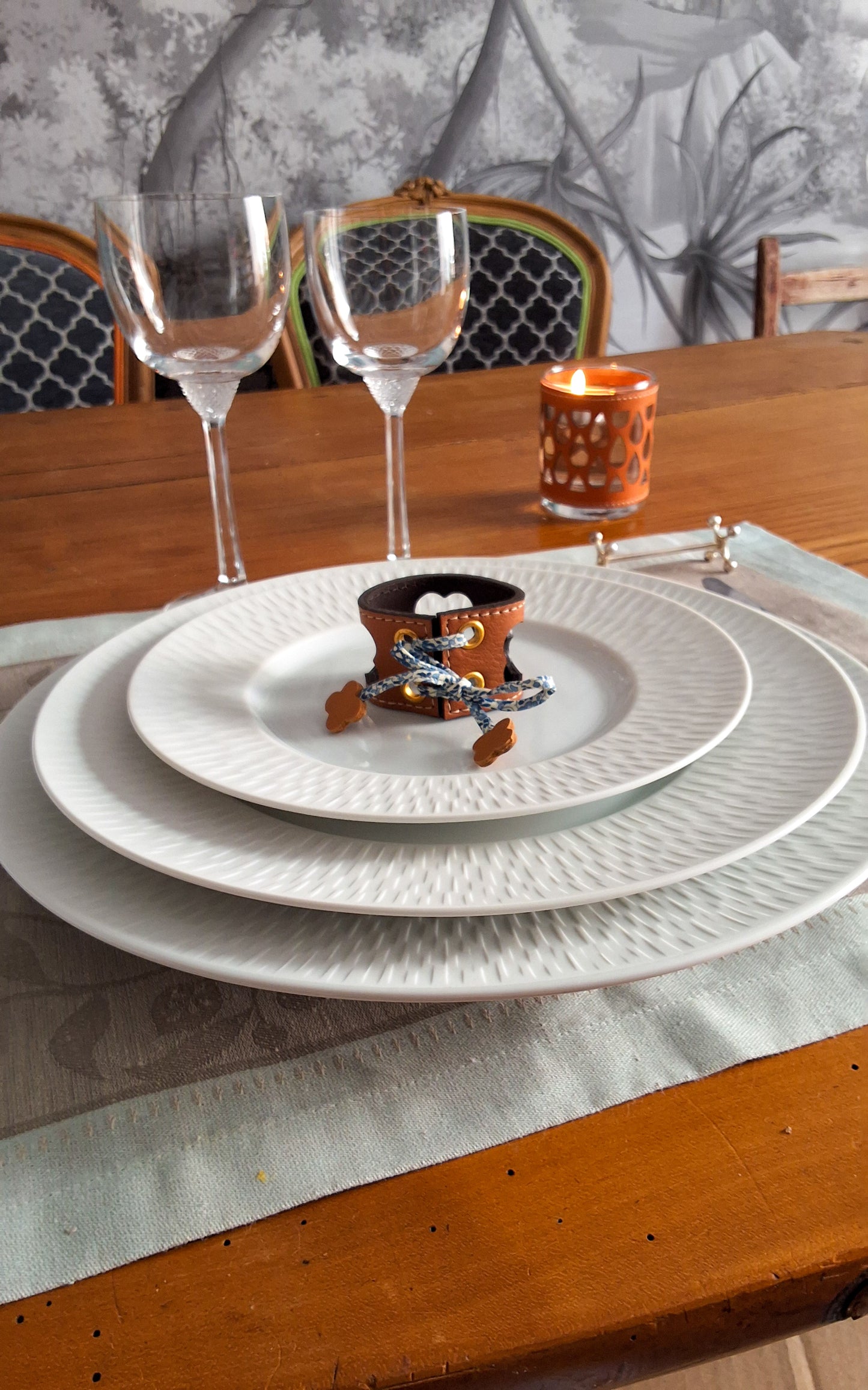 Table élégamment dressée avec assiettes superposées avec au centre un rond de serviette en cuir marron avec motif trèfle ajouré, verres à vin et bougie.