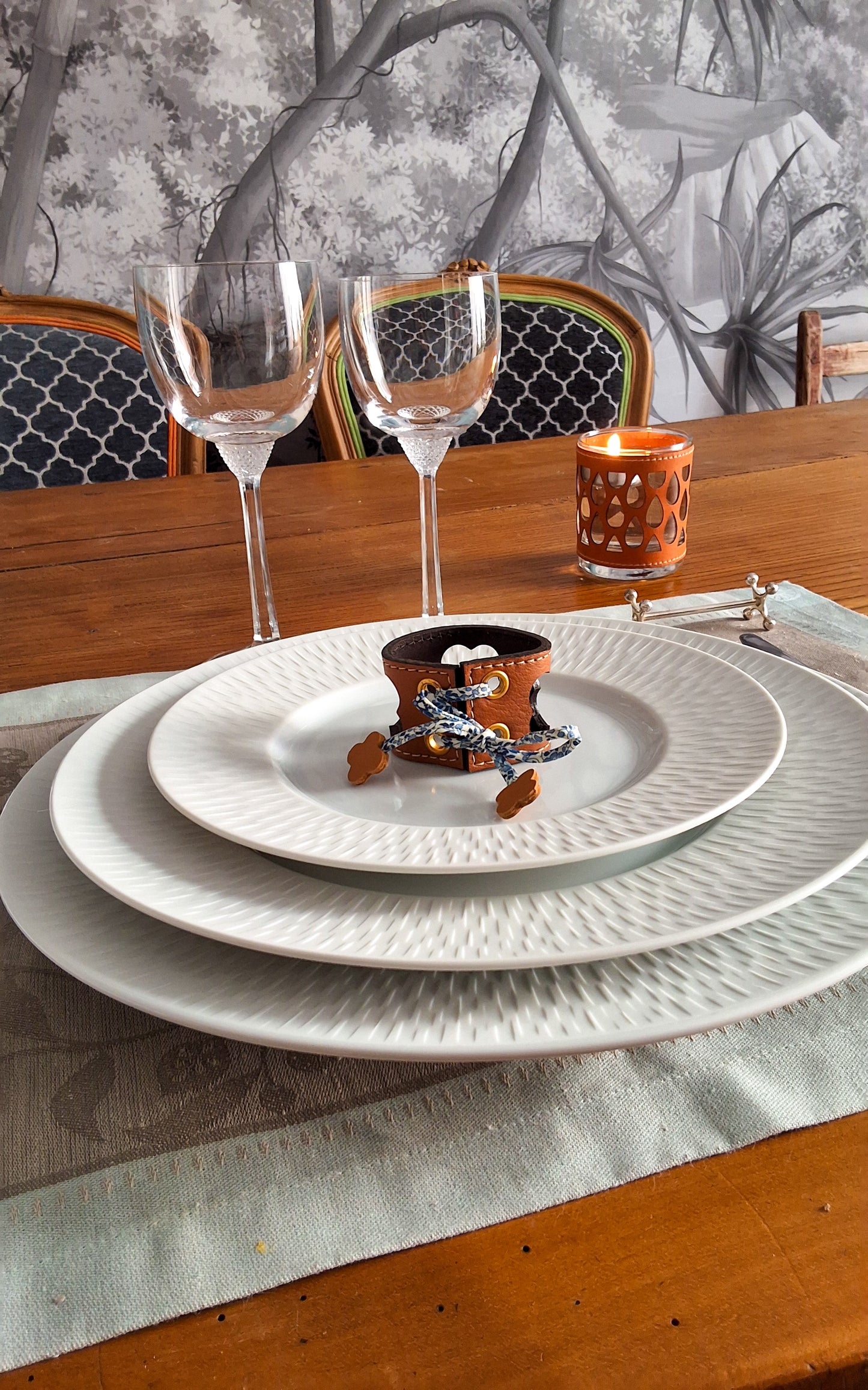 Table élégamment dressée avec assiettes superposées avec en son centre un rond de serviette en cuir marron avec motif trèfle ajouré, verres à vin et bougie.