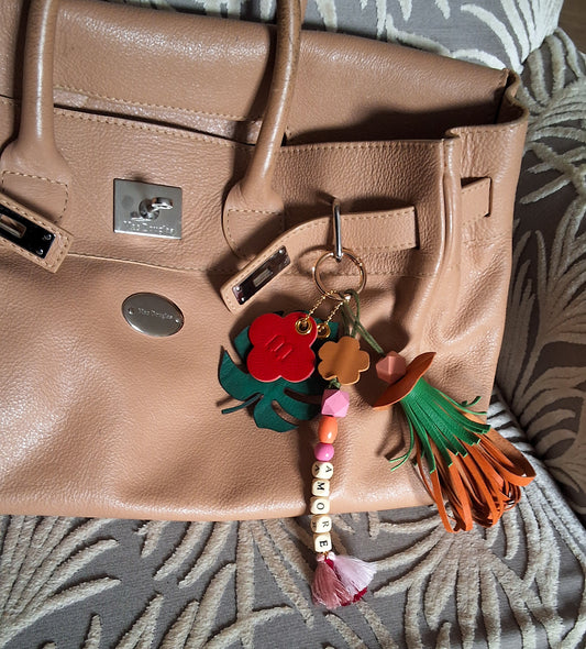 Bijou de sac avec quatre breloques en cuir fixées à un mousqueton doré, disposé sur un sac camel : un pompon orange et vert avec une perle de bois multifacette orange clair, une breloque en perles formant le mot « AMORE » avec trois petits pompons en fils de coton roses, une breloque en forme de feuille exotique verte avec une face en cuir façon crocodile et une face en cuir d’agneau vert sapin, et une breloque en forme de trèfle marquée M orange et rouge.