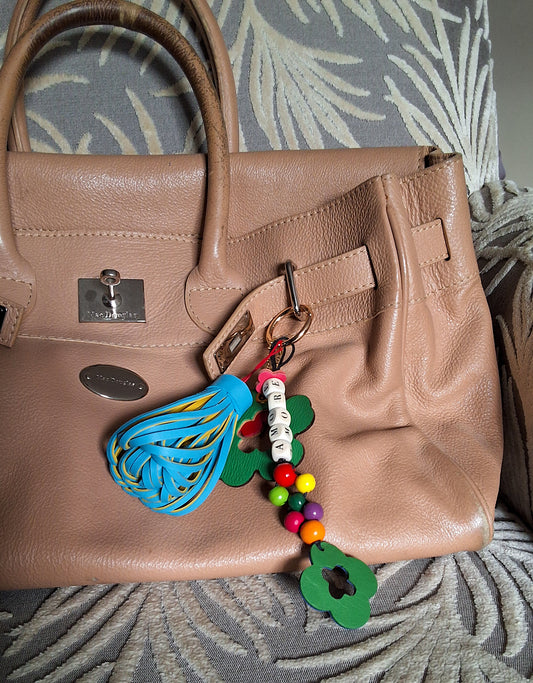 Bijou de sac en cuir et perles de bois multicolores avec anneau mousqueton doré, composé de quatre breloques, disposé sur un sac camel : un pompon bicolore bleu et jaune, une breloque en cuir vert et marron en forme de fleur évidée au centre, une breloque en cuir marron et rouge en forme de fleur marquée M, et une breloque en perles de bois formant le mot « AMORE » avec des perles rondes multicolores et un trèfle en cuir bleu et vert.