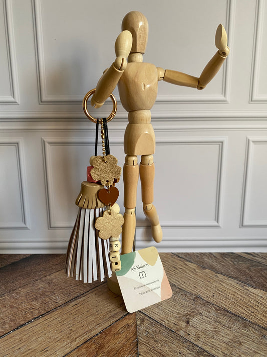 Bijou de sac avec trois breloques en cuir fixées à un mousqueton doré : un pompon mauve clair et beige avec deux perles en bois, une breloque en cuir en forme de guirlande dorée et rose dragée, et une breloque en perles de bois formant le mot « BRILLANTE ».