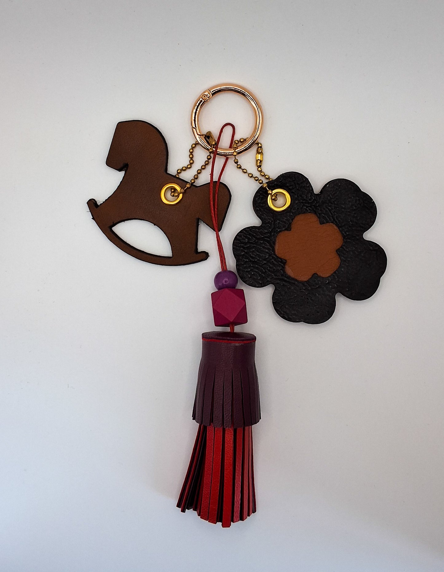 Bijou de sac avec trois breloques en cuir fixées à un mousqueton doré : un pompon bordeaux et rouge avec deux perles en bois et un cordon rouge, une breloque en cuir en forme de cheval à bascule marron et noir, et une breloque en forme de fleur avec un cœur marron marquée du M au dos.