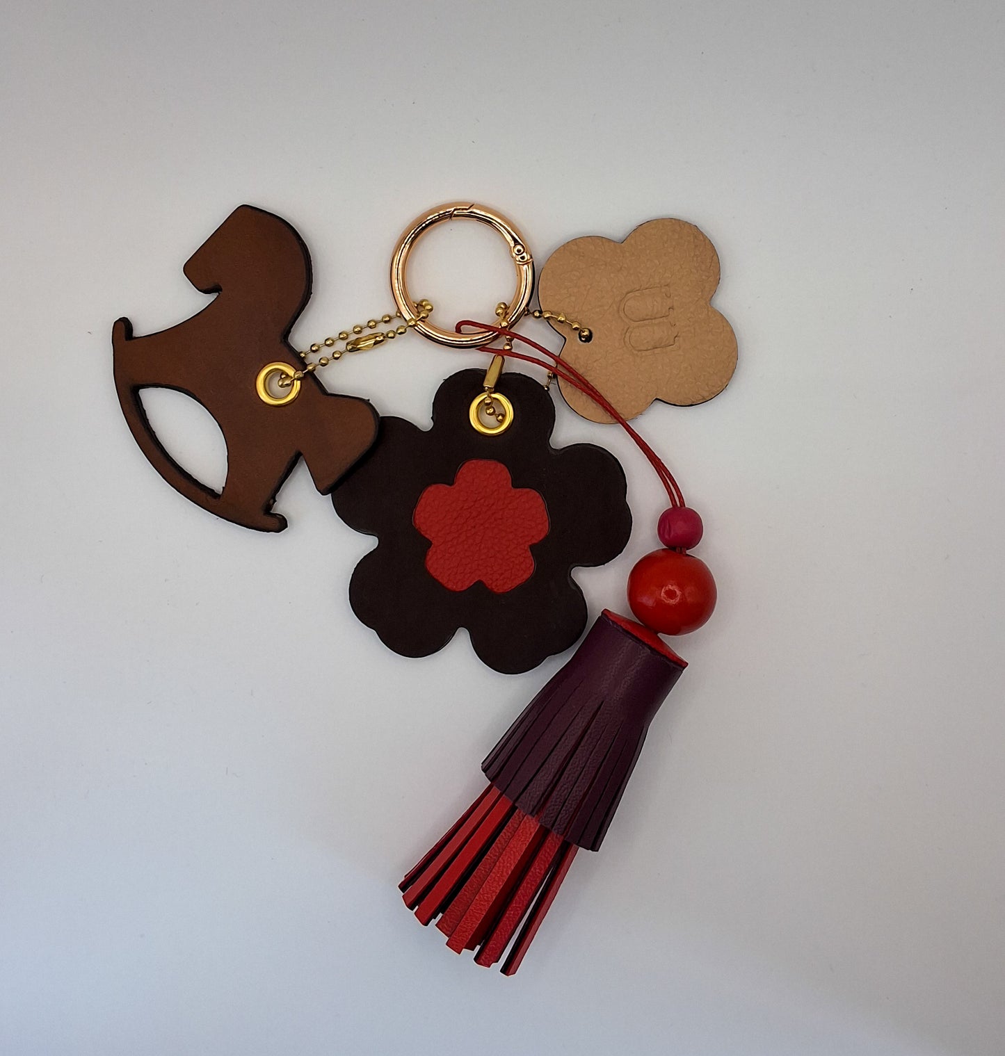 Bijou de sac avec quatre breloques en cuir fixées à un mousqueton doré : un pompon bordeaux et rouge avec deux perles en bois et un cordon rouge, une breloque en cuir en forme de cheval à bascule marron et noir, une breloque en cuir en forme de fleur avec un cœur rouge, et une breloque en cuir en forme de trèfle beige et marron marquée du M de la marque.