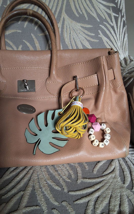 Bijou de sac avec trois breloques en cuir fixées à un mousqueton doré, disposé sur un sac camel : un pompon jaune et vert amande ceinturé d’un galon en gros grain marqué « amour toujours », une breloque en perles formant le mot « ÉTERNITÉ » avec deux perles en bois multifacette roses, et une breloque en forme de feuille exotique vert amande.