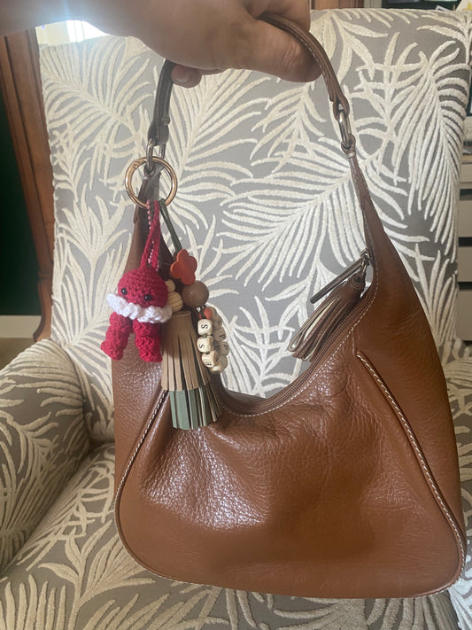 Bijou de sac en cuir avec quatre breloques fixées à un mousqueton doré ovale : un pompon beige et vert tendre avec une perle en bois, une breloque en perles de bois formant le mot « HAPPINESS », une breloque ronde en cuir marron ajourée d’un cœur, et une breloque en crochet faite à la main de couleur rouge.