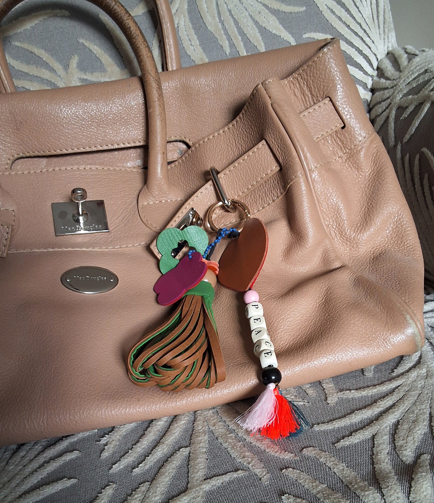 Bijou de sac en cuir et perles multicolores avec anneau doré, composé de quatre breloques, disposé sur un sac camel : un pompon vert et marron avec perle et petit trèfle orange, un trèfle vert et bleu évidé, un trèfle rose et rouge marqué M, et une breloque en perles formant « PEACE » avec trois pompons et un cœur rouge et marron.