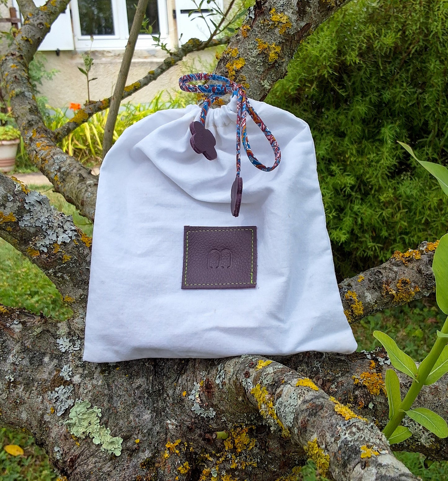 Pochon de rangement blanc avec au centre une pièce de cuir carré violette embossée avec le M de la marque et des cordons liberty violet disposé sur un arbre dans un jardin.