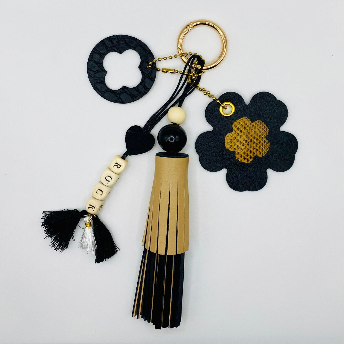 Bijou de sac avec quatre breloques en cuir fixées à un mousqueton doré : un pompon noir et beige avec deux perles en bois, une breloque ronde en cuir noir effet crocodile avec un trèfle, une breloque en forme de fleur avec un cœur en peau de serpent et un M au dos, et une breloque en perles de bois formant le mot « ROCK ».