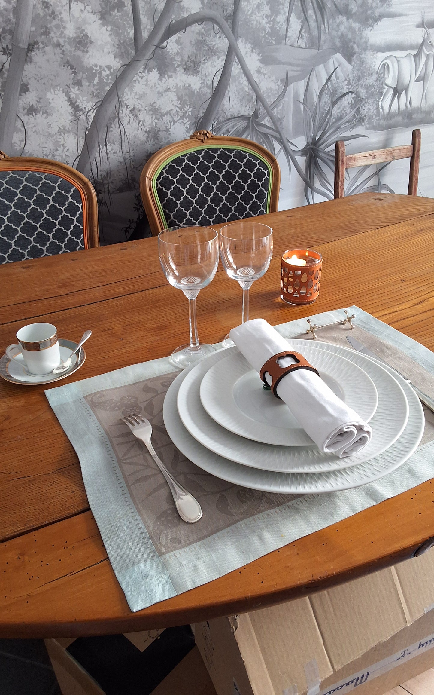 Table élégamment dressée avec assiettes superposées, serviette blanche roulée maintenue par un rond de serviette en cuir marron avec motif trèfle ajouré, verres à vin et bougie.
