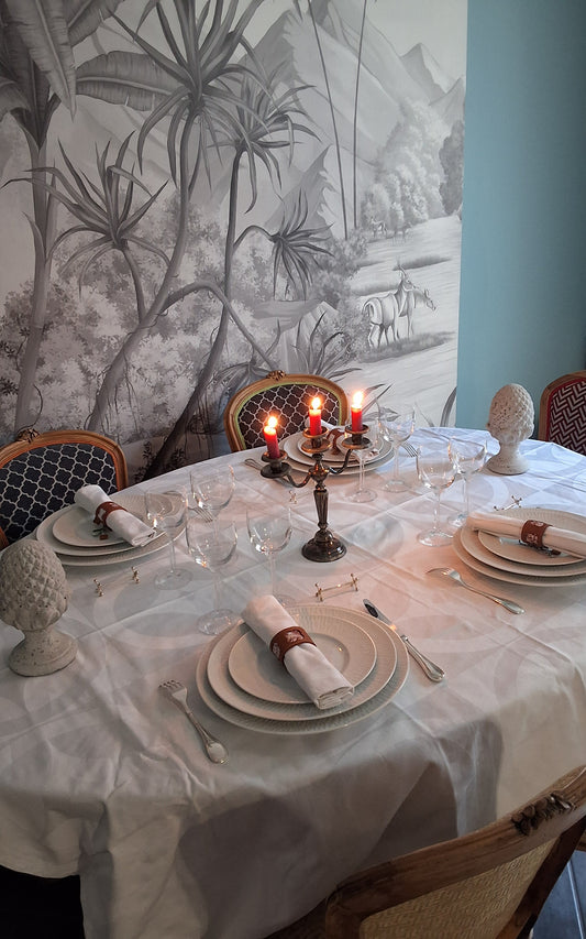 Table élégamment dressée avec assiettes superposées, serviettes blanches roulées maintenues par un rond de serviette en cuir marron avec motif embossé, verres à vin et chandelier à trois bougies rouges allumées au centre.
