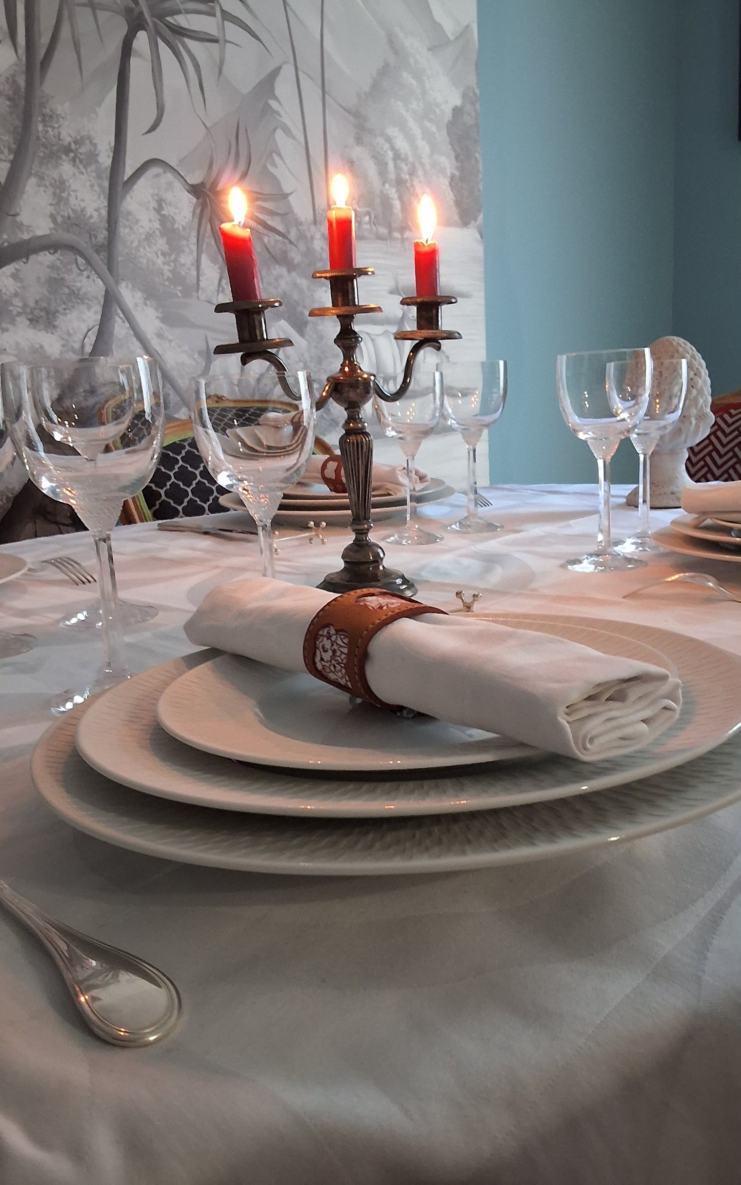Table élégamment dressée avec assiettes superposées, serviette blanche roulée maintenue par un rond de serviette en cuir marron avec motif embossé, verres à vin et chandelier à trois bougies rouges allumées au centre.