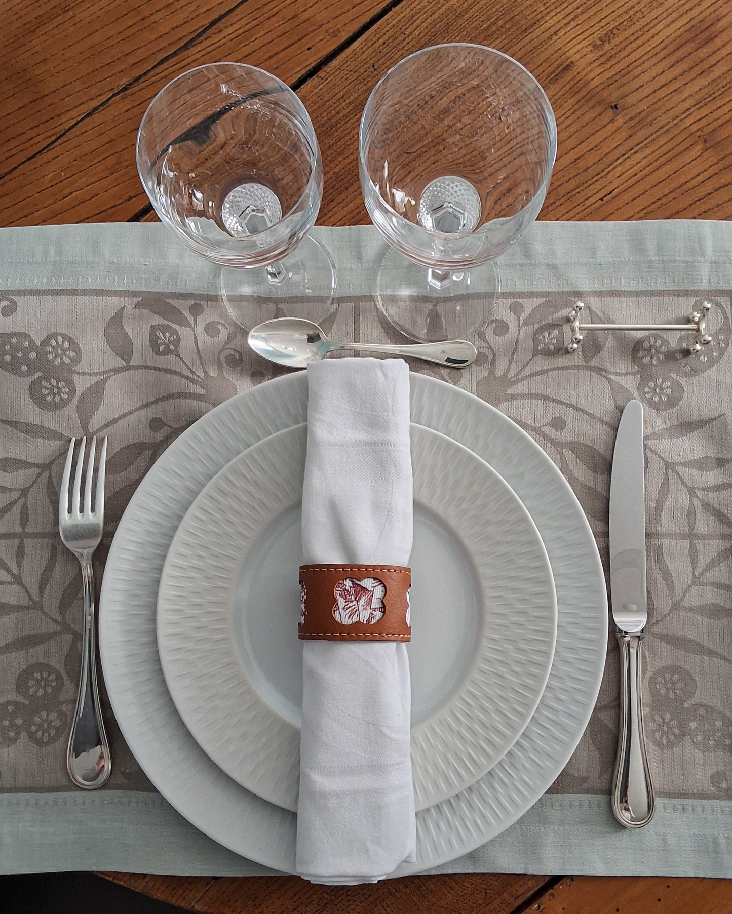 Serviette blanche roulée maintenue par un rond de serviette en cuir marron avec motif embossé sur deux assiettes superposées, argenterie élégante sur un set de table lui-même disposé sur une table en bois.