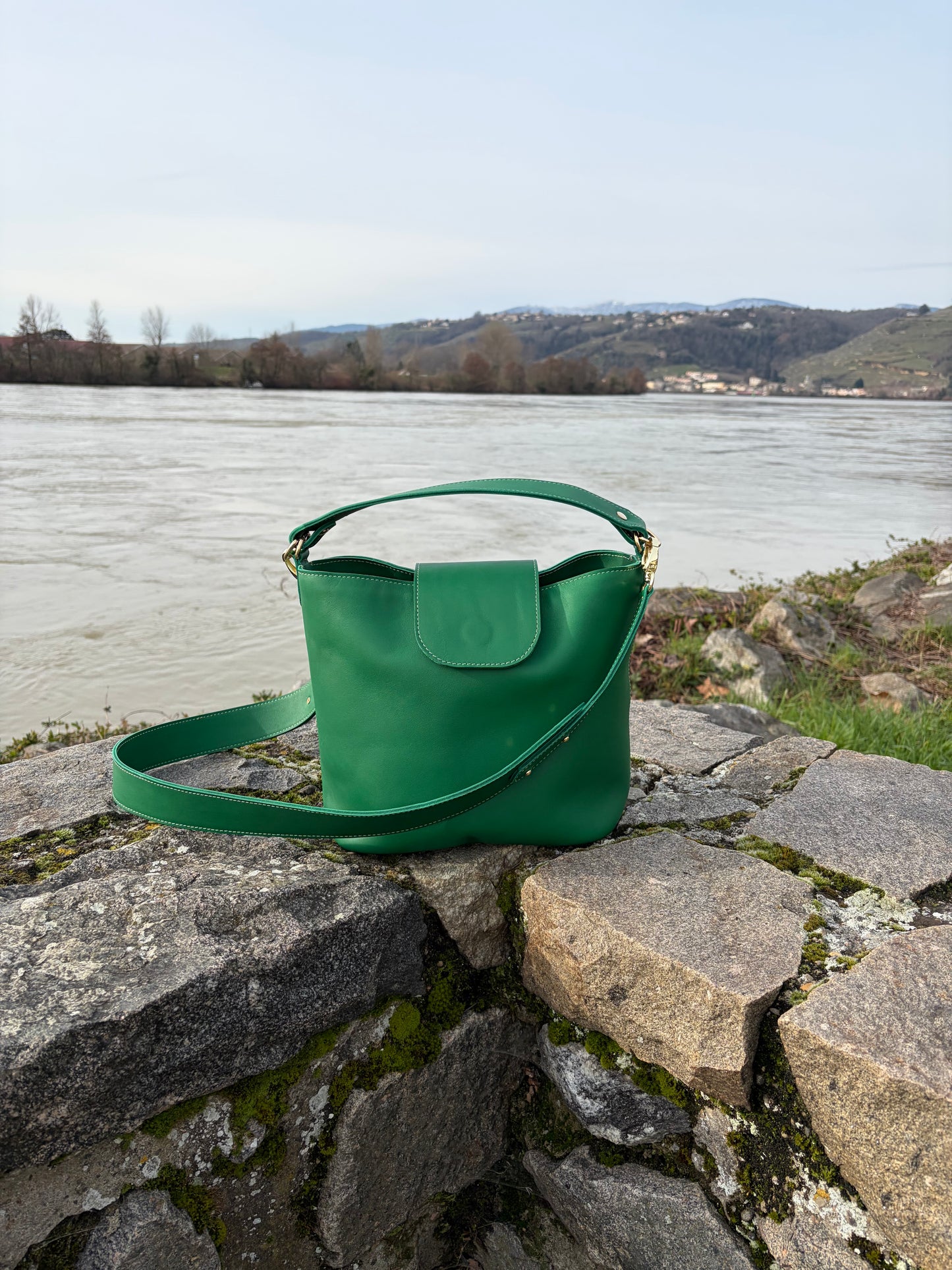 photo du sac seau garance en cuir de veau vert posé sur un muret en pierre le long du fleuve Rhône