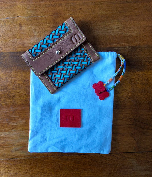Porte cartes camel surpiqûres jaunes et motif tressage bleu turquoise et pochon de rangement blanc avec en bas au centre une pièce de cuir carré rouge embossée avec le M de la marque et des cordons liberty rouge disposés à plat sur une table en bois.