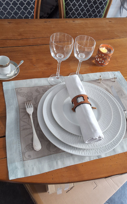 Table élégamment dressée avec assiettes superposées, serviette blanche roulée maintenue par un rond de serviette en cuir marron avec motif trèfle ajouré, verres à vin et bougie.