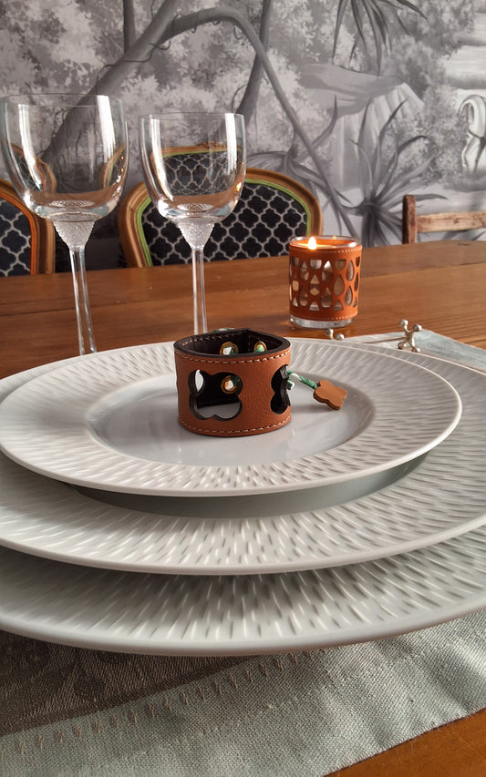 Table élégamment dressée avec assiettes  et au centre un rond de serviette en cuir marron avec motif trèfle ajouré, verres à vin et bougie.