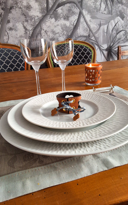 Table élégamment dressée avec assiettes superposées avec en son centre un rond de serviette en cuir marron avec motif trèfle ajouré, verres à vin et bougie.