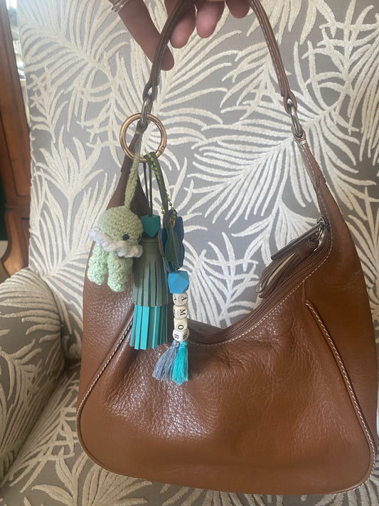 Bijou de sac en cuir avec quatre breloques fixées à un mousqueton doré ovale : un pompon azur et vert tendre avec une perle en bois, une breloque en perles de bois formant le mot « AMOR », une breloque florale en cuir marron et crocodile bleu marquée de la lettre M, et une breloque en crochet faite à la main de couleur vert amande.