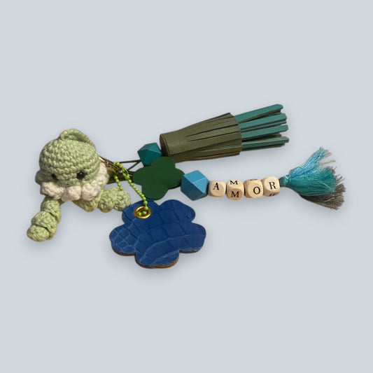 Bijou de sac en cuir avec quatre breloques fixées à un mousqueton doré ovale : un pompon azur et vert tendre avec une perle en bois, une breloque en perles de bois formant le mot « AMOR », une breloque florale en cuir marron et crocodile bleu marquée de la lettre M, et une breloque en crochet faite à la main de couleur vert amande.