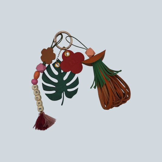 Bijou de sac avec quatre breloques en cuir fixées à un mousqueton doré : un pompon orange et vert avec une perle de bois multifacette orange clair, une breloque en perles formant le mot « AMORE » avec trois petits pompons en fils de coton roses, une breloque en forme de feuille exotique verte avec une face en cuir façon crocodile et une face en cuir d’agneau vert sapin, et une breloque en forme de trèfle marquée M orange et rouge.