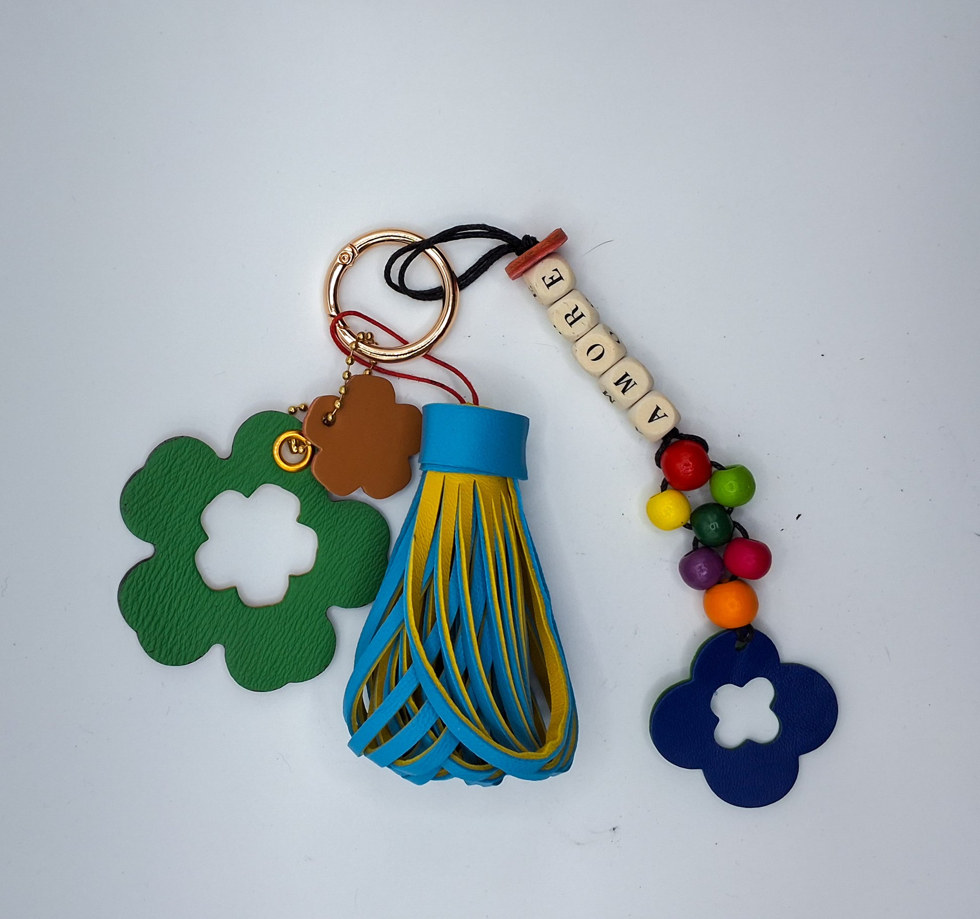 Bijou de sac en cuir et perles de bois multicolores avec anneau mousqueton doré, composé de quatre breloques : un pompon bicolore bleu et jaune, une breloque en cuir vert et marron en forme de fleur évidée au centre, une breloque en cuir marron et rouge en forme de fleur marquée M, et une breloque en perles de bois formant le mot « AMORE » avec des perles rondes multicolores et un trèfle en cuir bleu et vert.