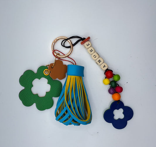 Bijou de sac en cuir et perles de bois multicolores avec anneau mousqueton doré, composé de quatre breloques : un pompon bicolore bleu et jaune, une breloque en cuir vert et marron en forme de fleur évidée au centre, une breloque en cuir marron et rouge en forme de fleur marquée M, et une breloque en perles de bois formant le mot « AMORE » avec des perles rondes multicolores et un trèfle en cuir bleu et vert.