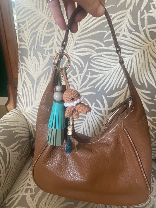 Bijou de sac en cuir avec quatre breloques fixées à un mousqueton doré ovale : un pompon azur et vert tendre avec une perle en bois, une breloque en perles de bois formant le mot « AMOUR », une breloque en cuir en forme de fleur marquée de la lettre M, et une breloque en crochet faite à la main de couleur marron.