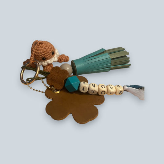 Bijou de sac en cuir avec quatre breloques fixées à un mousqueton doré ovale : un pompon azur et vert tendre avec une perle en bois, une breloque en perles de bois formant le mot « AMOUR », une breloque en cuir en forme de fleur marquée de la lettre M, et une breloque en crochet faite à la main de couleur marron.