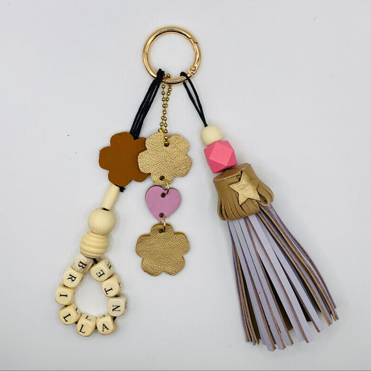 Bijou de sac avec trois breloques en cuir fixées à un mousqueton doré : un pompon mauve clair et beige avec deux perles en bois, une breloque en cuir en forme de guirlande dorée et rose dragée, et une breloque en perles de bois formant le mot « BRILLANTE ».