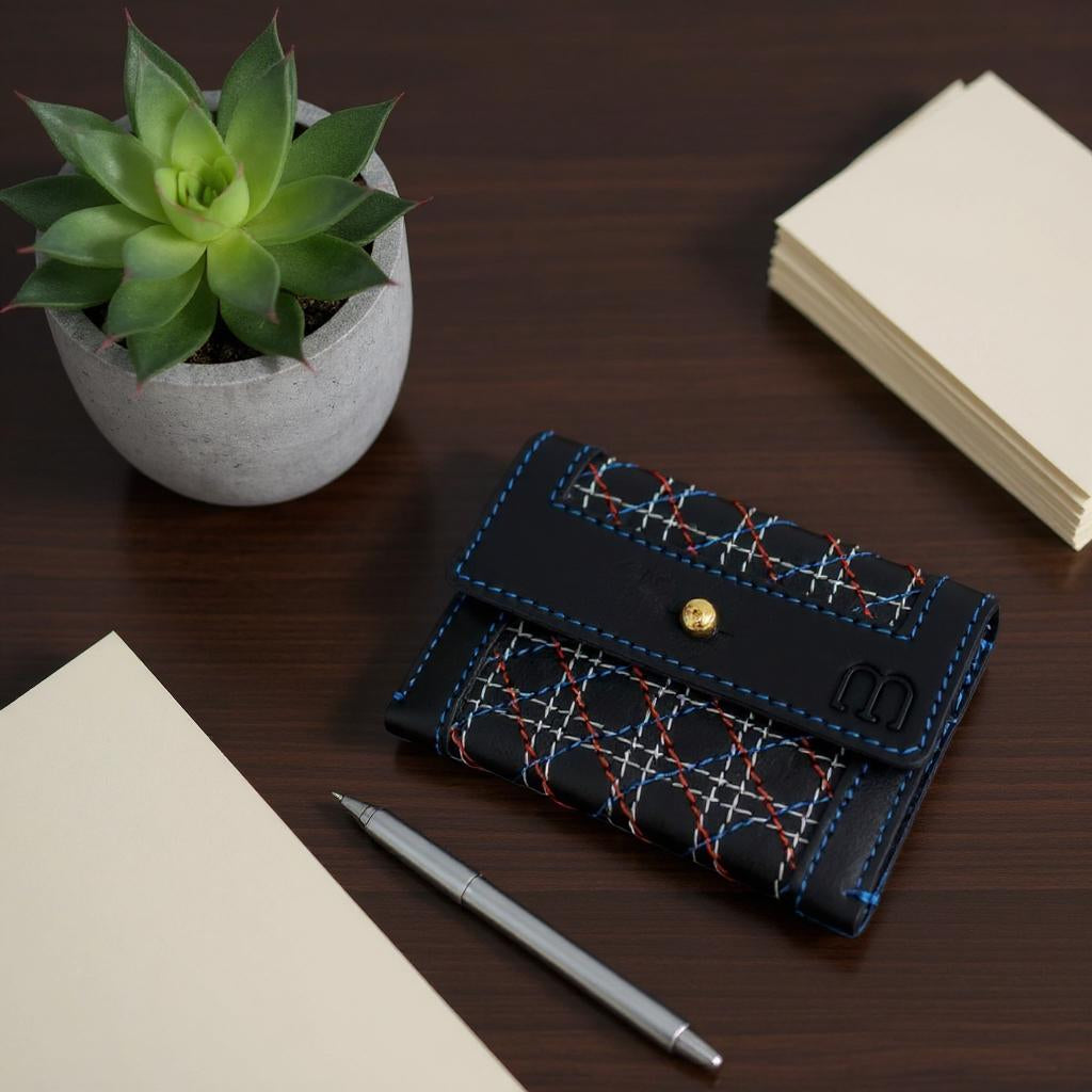 Porte cartes en cuir noir avec motifs de surpiqûres croisées rouges, blanches et bleues, fermé par un bouton doré, posé sur un bureau en bois foncé. À côté, un stylo argenté, une pile de feuilles et une de cartes beige, et un petit pot en béton contenant une plante succulente verte.