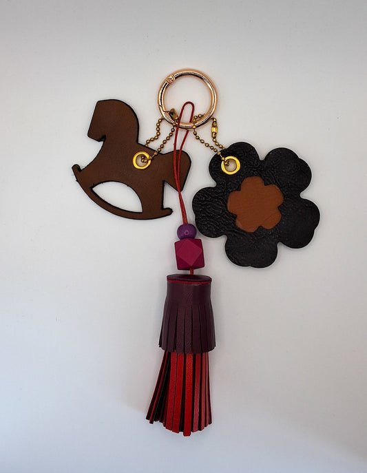 Bijou de sac avec trois breloques en cuir fixées à un mousqueton doré : un pompon bordeaux et rouge avec deux perles en bois et un cordon rouge, une breloque en cuir en forme de cheval à bascule marron et noir, et une breloque en forme de fleur avec un cœur marron marquée du M au dos.