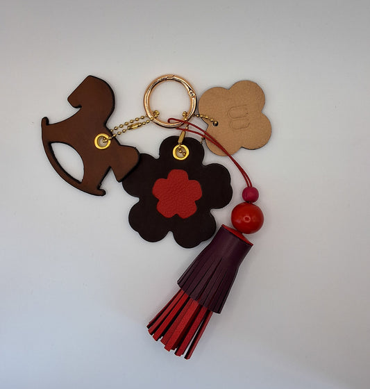 Bijou de sac avec quatre breloques en cuir fixées à un mousqueton doré : un pompon bordeaux et rouge avec deux perles en bois et un cordon rouge, une breloque en cuir en forme de cheval à bascule marron et noir, une breloque en cuir en forme de fleur avec un cœur rouge, et une breloque en cuir en forme de trèfle beige et marron marquée du M de la marque.