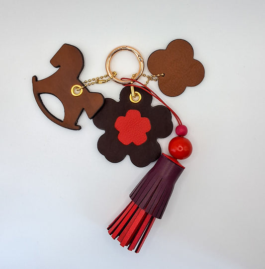Bijou de sac avec quatre breloques en cuir fixées à un mousqueton doré : un pompon bordeaux et rouge avec deux perles en bois et un cordon rouge, une breloque en cuir en forme de cheval à bascule marron et noir, une breloque en cuir en forme de fleur avec un cœur rouge, et une breloque en cuir en forme de trèfle beige et marron marquée du M de la marque.