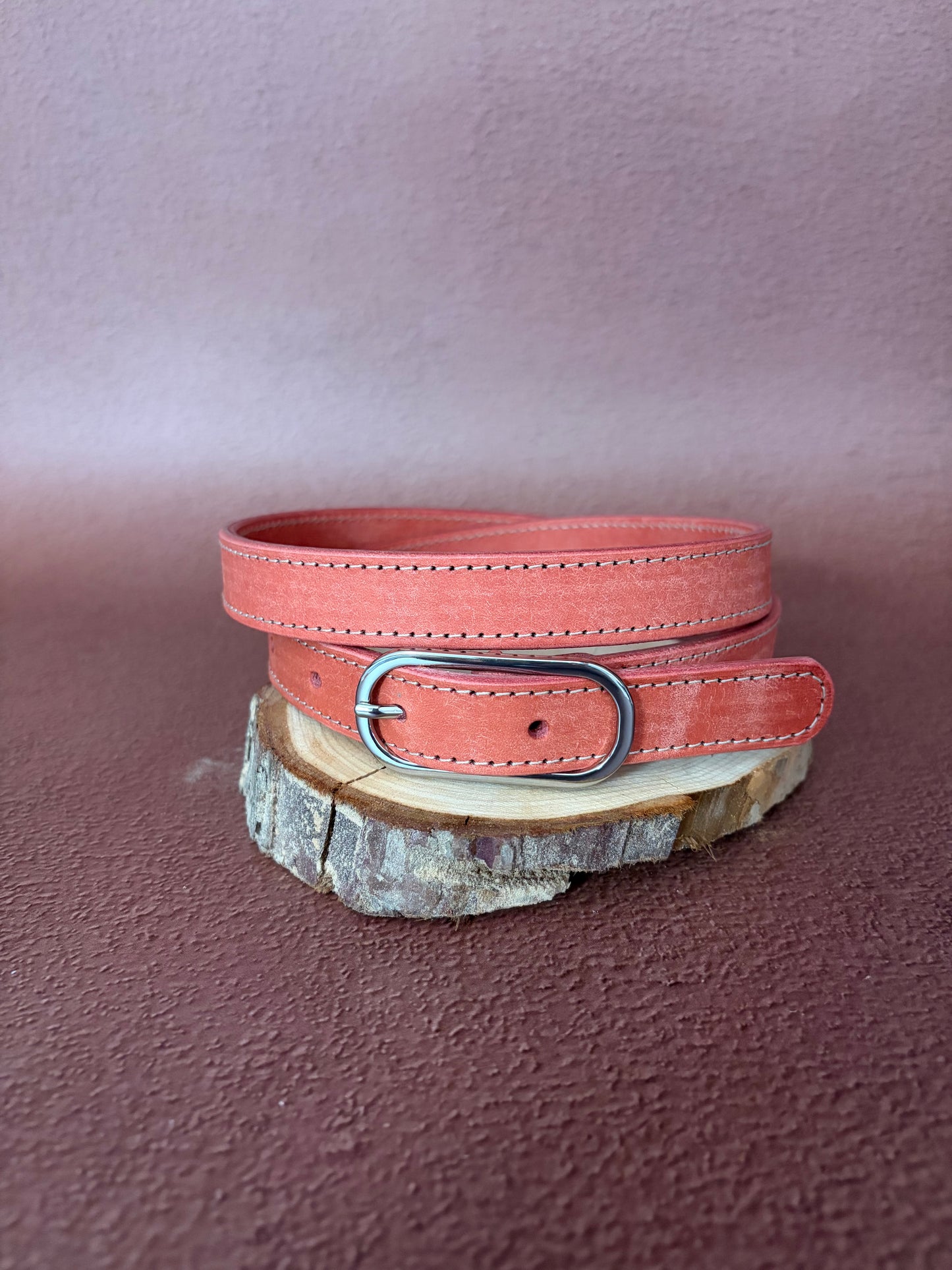 une ceinture en cuir tannage végétal rose avec une boucle ovale en acier est posée sur une tranche de bois le tout sur un fond ocre texturé
