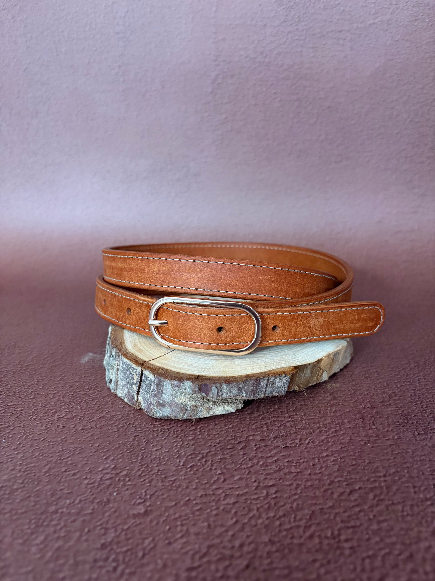 une ceinture en cuir fauve au tannage végétal est posée sur une tranche de bois le tout sur un fond ocre