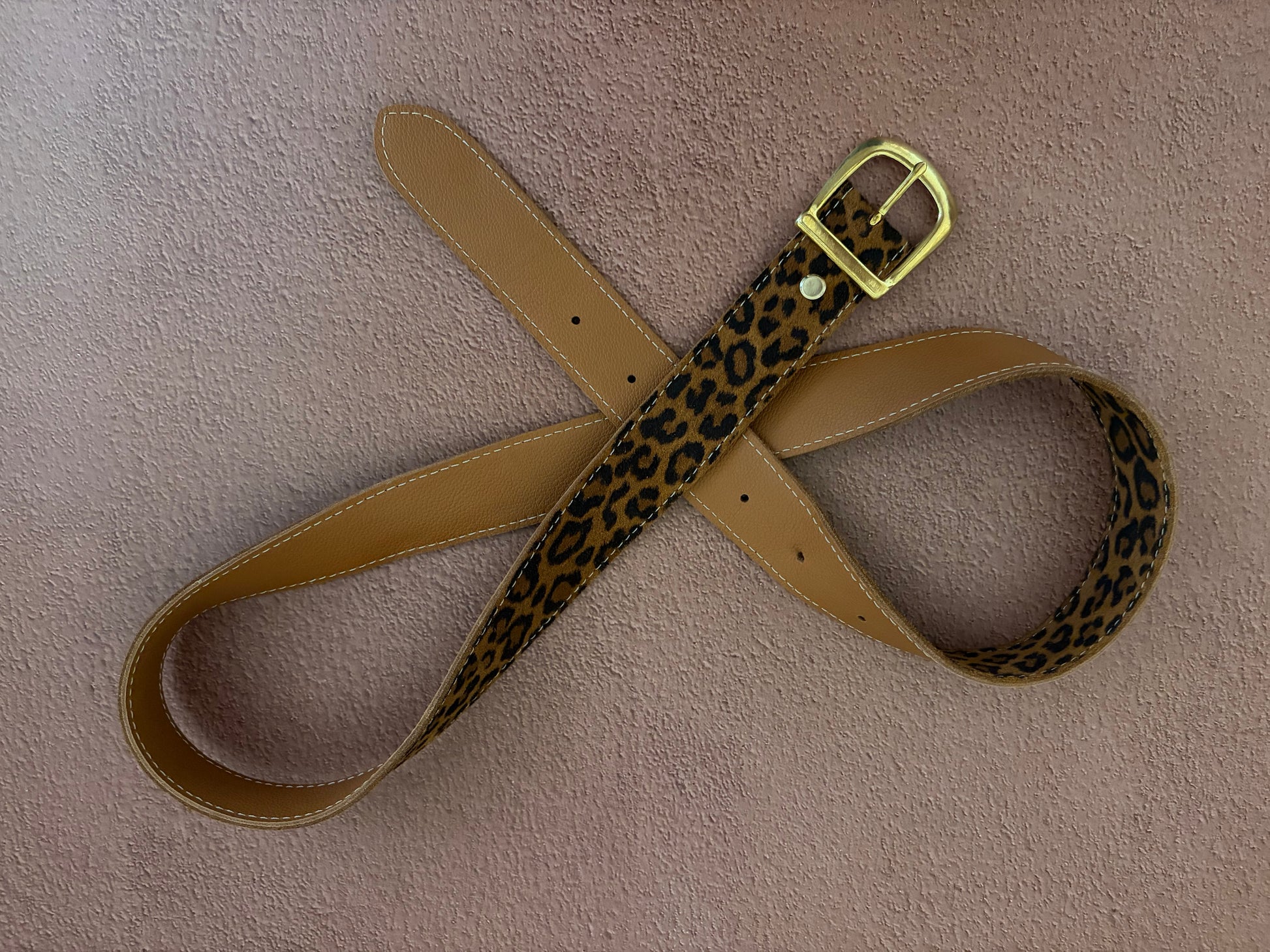 la ceinture en cuir fantaisie léopard est présentée sur un fond texturé ocre. la ceinture est positionnée en boucles afin de montrer le recto en cuir léopard et le verso en cuir marron grainé. 