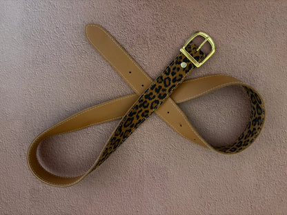 la ceinture en cuir fantaisie léopard est présentée sur un fond texturé ocre. la ceinture est positionnée en boucles afin de montrer le recto en cuir léopard et le verso en cuir marron grainé. 