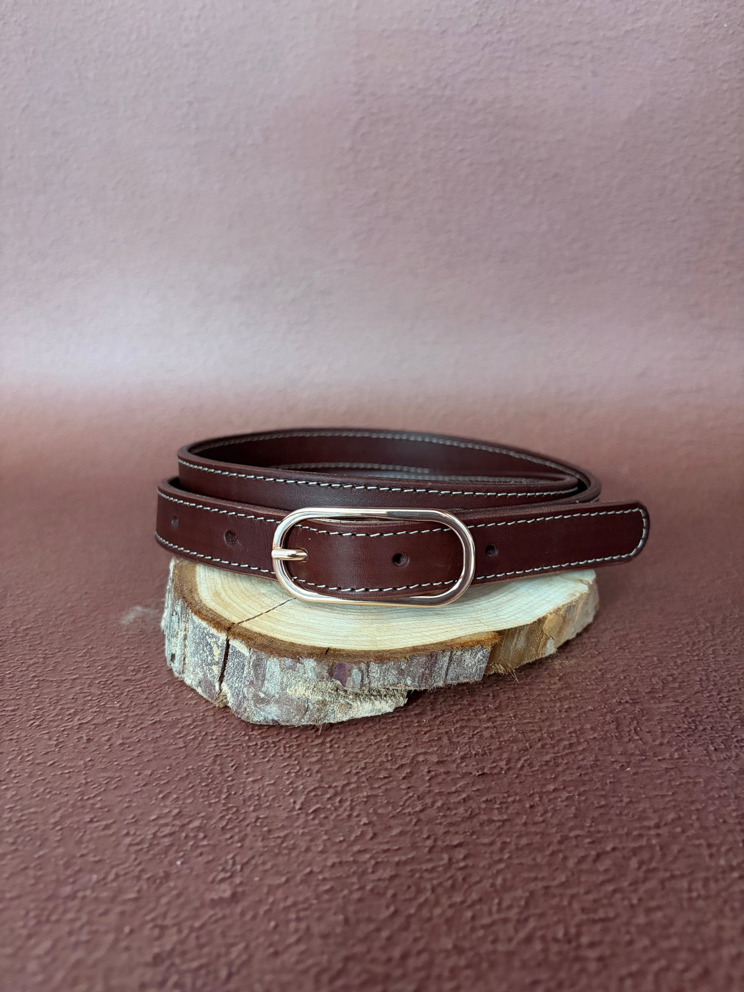 une ceinture en cuir naturel marron tannage végétal avec une boucle ovale dorée est posée sur une tranche de bois le tout sur un fond ocre