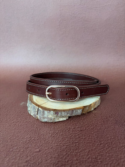 une ceinture en cuir naturel marron tannage végétal avec une boucle ovale dorée est posée sur une tranche de bois le tout sur un fond ocre