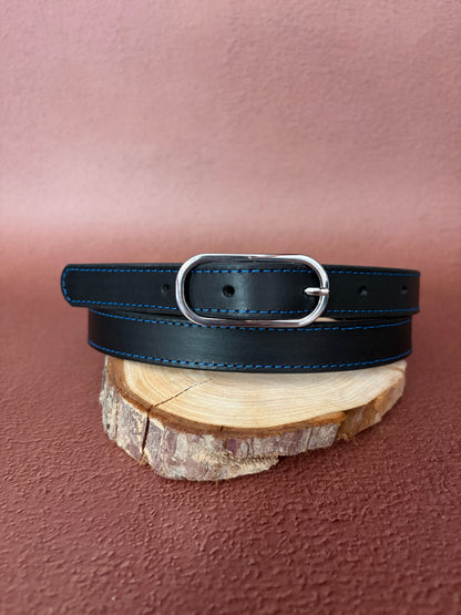 une ceinture fine en cuir noire et piqures bleu avec une boucle ovale en acier est posée sur une tranche de bois le tout sur un fond ocre
