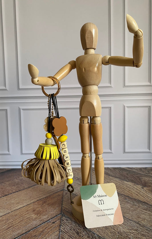 Bijou de sac avec trois breloques en cuir fixées à un mousqueton doré : un pompon jaune et beige avec deux perles en bois, une breloque en cuir en forme de guirlande dorée et noire, et une breloque en perles de bois formant le mot « CHANCE ».
