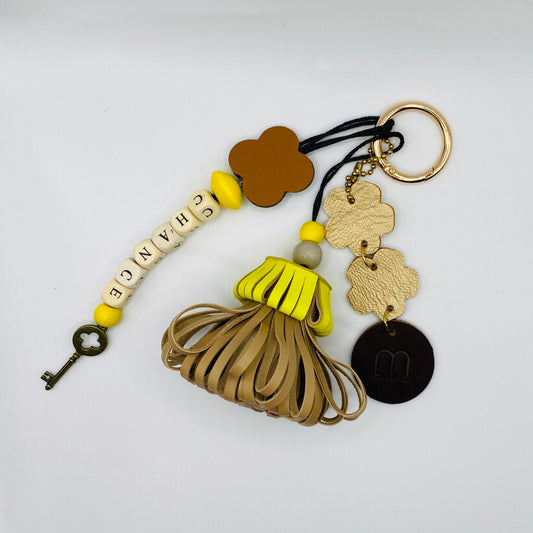 Bijou de sac avec trois breloques en cuir fixées à un mousqueton doré : un pompon jaune et beige avec deux perles en bois, une breloque en cuir en forme de guirlande dorée et noire, et une breloque en perles de bois formant le mot « CHANCE ».