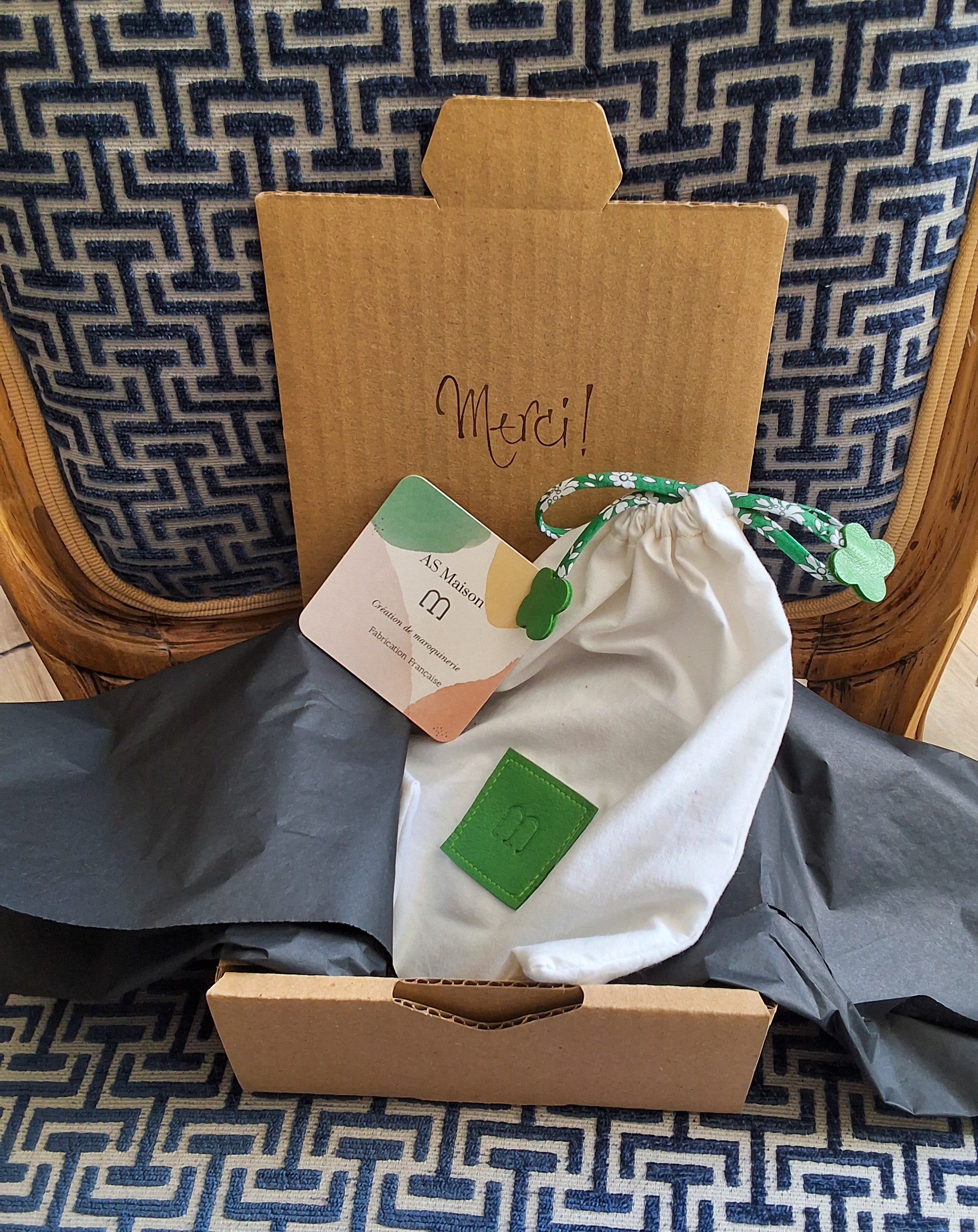 Pochon de rangement blanc et vert disposé dans un papier de soie gris foncé lui même disposé dans une boîte en carton avec le mot Merci sur une chaise.