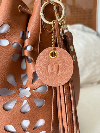 vue en gros plan de la pièce ronde en cuir qui porte la marque. en arrière plan on voir le pompon en cuir ainsi que le détail du motif perforé du sac. 