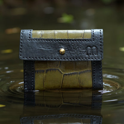 Porte cartes noir et vert kaki croco et sigle M embossé à droite comme en lévitation sur un lac se reflétant dans l'eau.