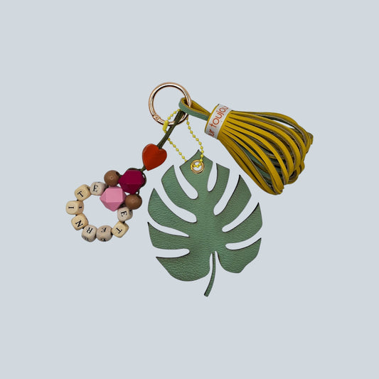 Bijou de sac avec trois breloques en cuir fixées à un mousqueton doré, disposé sur un sac camel : un pompon jaune et vert amande ceinturé d’un galon en gros grain marqué « amour toujours », une breloque en perles formant le mot « ÉTERNITÉ » avec deux perles en bois multifacette roses, et une breloque en forme de feuille exotique vert amande.