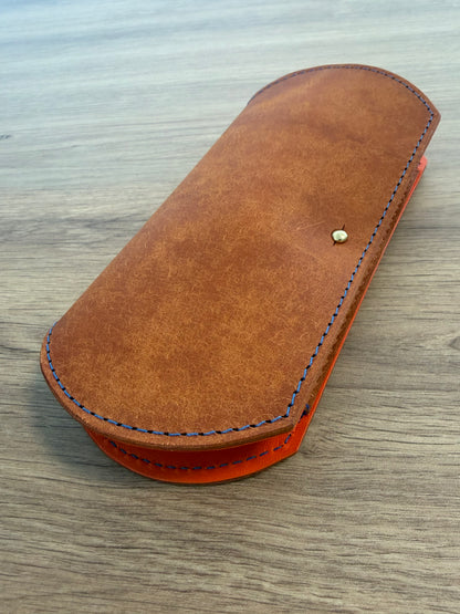 étui à lunette plat fermé réalisé en cuir au tannage végétal avec les surpiqures leur et un fermoir doré le tout posé sur une table en bois 