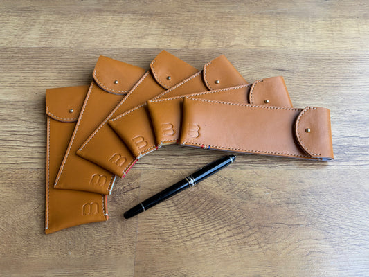 6 étuis à crayons en cuir marron clair glacé sont disposés en éventail sur un parquet ancien.