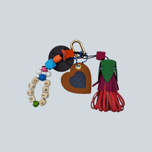 Bijou de sac en cuir avec quatre breloques fixées à un mousqueton doré ovale : un pompon tricolore orange, rose et vert avec une perle en bois multifacette, une breloque en perles formant le mot « GOOD TIME » avec des perles multicolores, une breloque en forme de cœur en cuir et peau de serpent d’eau, et une breloque ronde en cuir marron marquée M.
